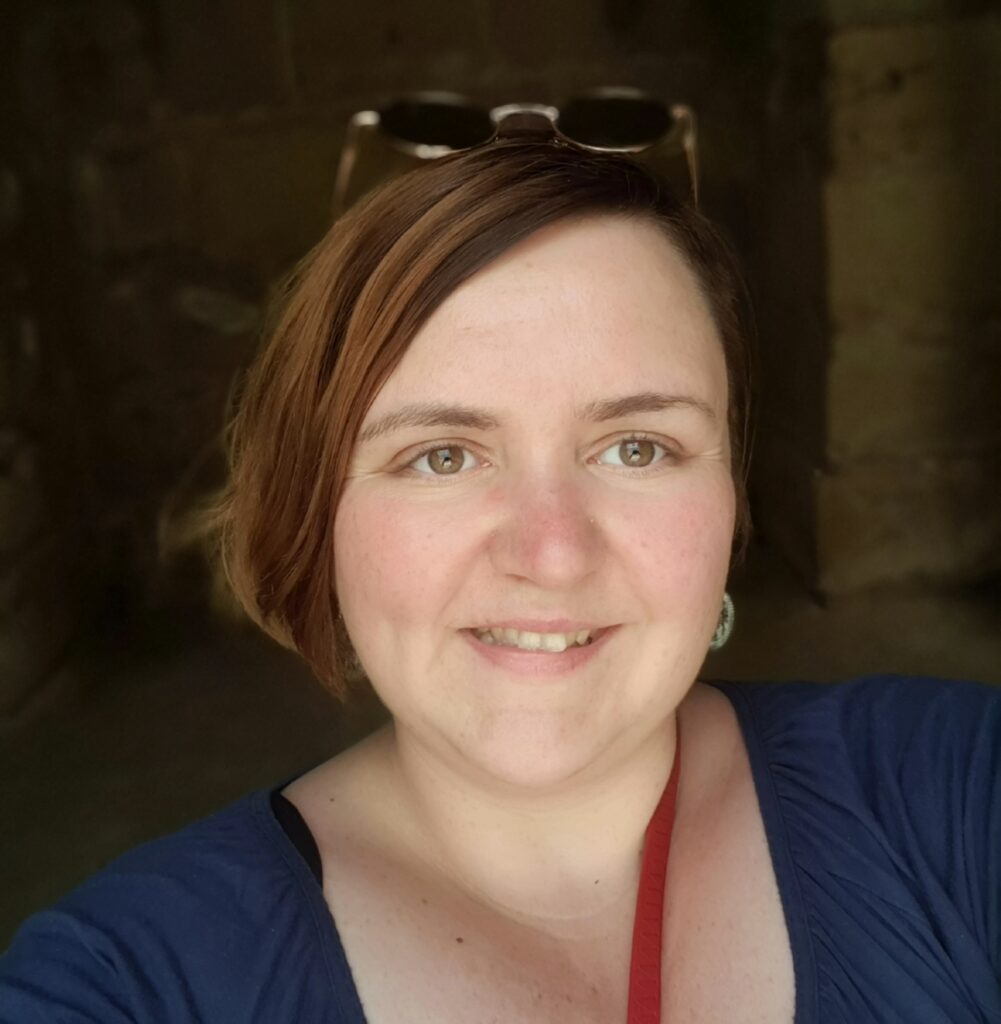 Foto einer Frau mit kinnblangem, braunem Haar und einem blauen Shirt, die etwa bis zu den Schultern zu sehen ist. Sie lächelt in die Kamera und hat eine Sonnenbrille auf die Stirn geschoben.